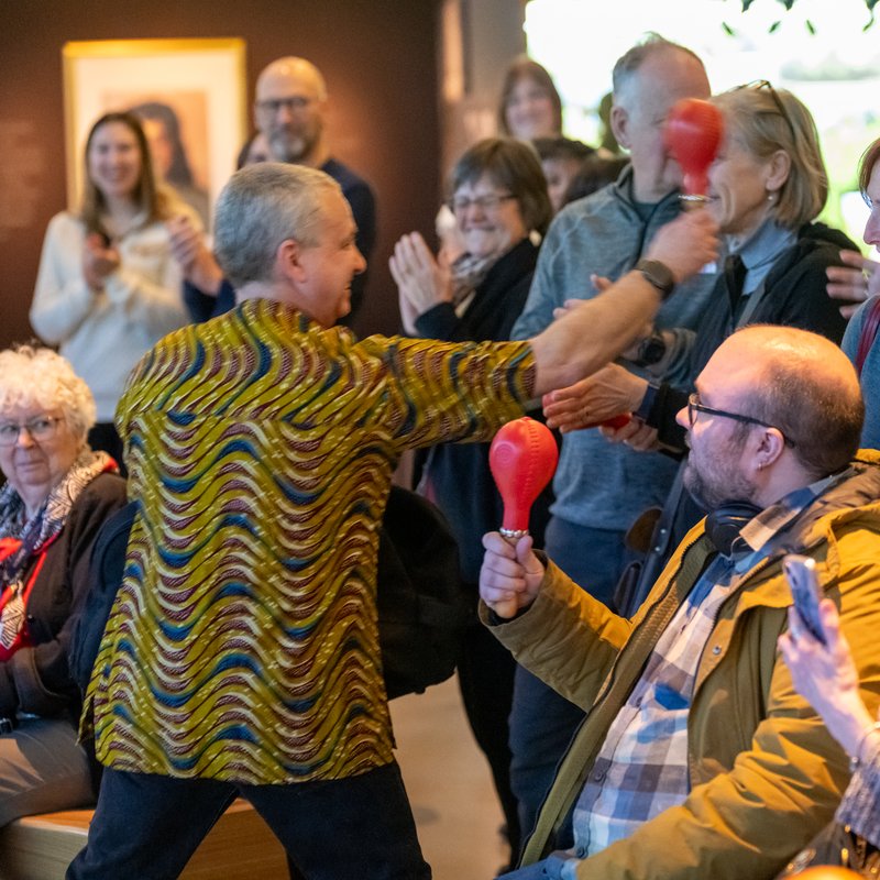 A Drum Cafe facilitator in a colourful shirt getting a smiling audience playing shakers