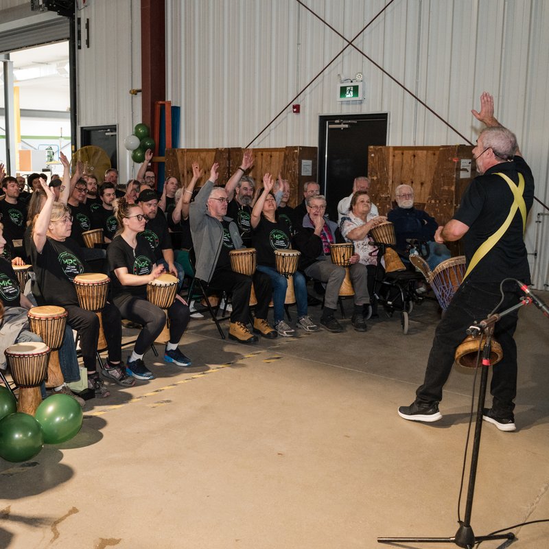A large group seated at hand drums raising their arms with a Drum Cafe facilitator